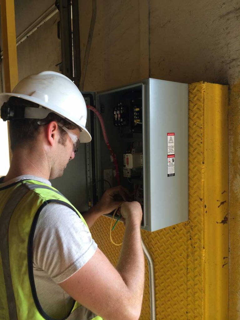 Man with hard hat, safety glasses, and reflective safety vest working on electrical service box.